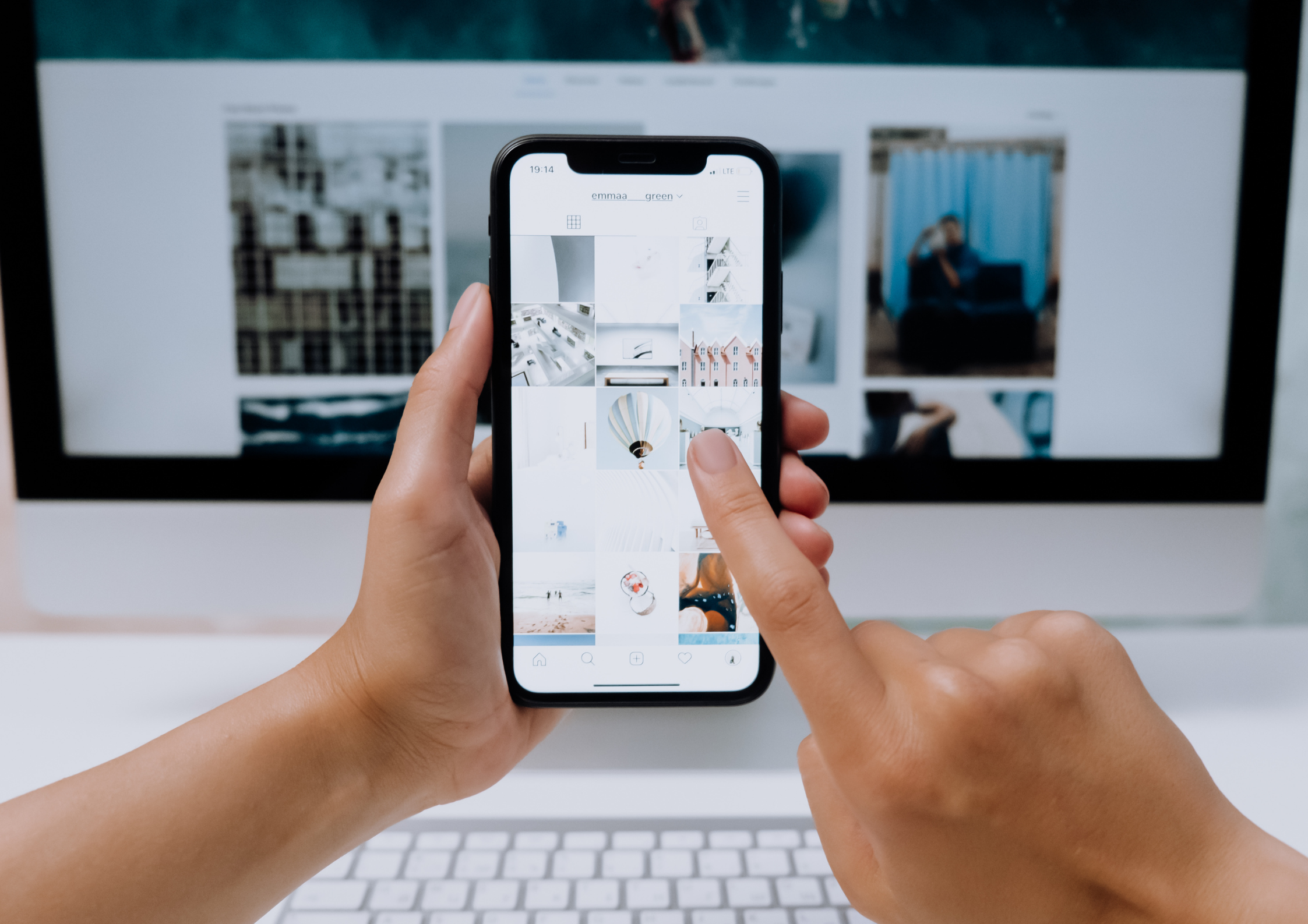 Person holding their phone in front of their computer, showing off their white themed instagram page